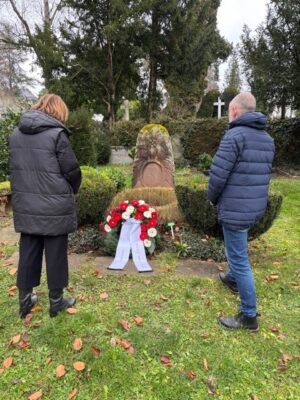 Anni Betz, Geschäftsführerin der Carlo-Schmid-Stiftung und Tobias Schmid, Enkel Carlo Schmids, stehen am Grab von Carlo Schmid, auf dem ein Kranz mit roten und weißen Blumen liegt.